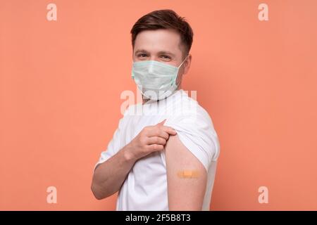 Homme caucasien en masque médical montrant son bras vacciné. Il a reçu un vaccin corona qui regarde la caméra. Prise de vue en studio Banque D'Images