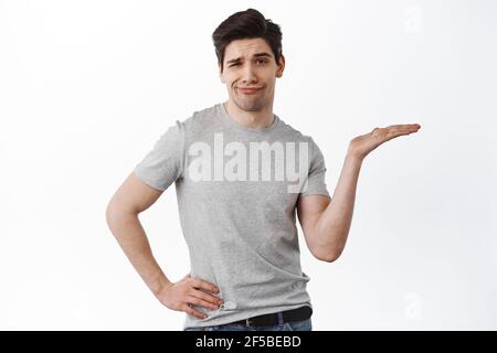 Homme adulte sceptique tenant à la main, montrer l'espace vide sur sa paume et regarder déçu, debout douteux sur fond blanc Banque D'Images
