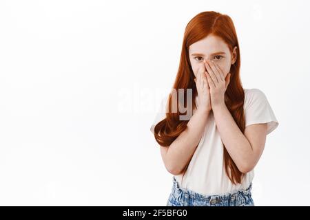 Une jeune fille à tête rouge couvre son visage et regarde surpris à l'appareil photo, à peine surpris. Un petit enfant se cachant derrière les mains et piquant à vous, debout Banque D'Images