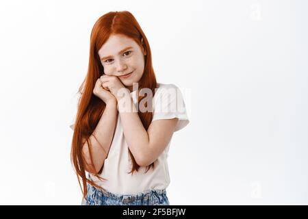 Jolie petite fille à tête rouge avec des taches de rousseur et de longs cheveux au gingembre, elle a l'air adorable et mignonne à l'appareil photo, tenant les mains près du visage dans une posture stupide, rêvant de jour Banque D'Images