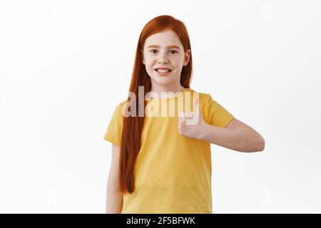 Portrait de fille de gingembre avec un sourire mignon et goûtant, montre le pouce vers le haut dans l'approbation, comme et d'accord, dire oui, donner une réponse positive, recommander quelque chose de bon Banque D'Images