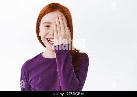 Joyeux redhead petite fille, enfant de gingembre cachant la moitié du visage derrière la main et rire, souriant gai, debout sur fond blanc Banque D'Images