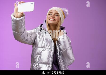 Heureuse ravie insouciante femme européenne charmante à cheveux justes en hiver argenté veste chapeau levant le smartphone horizontalement prendre selfie sourire mobile Banque D'Images