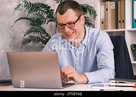 Joyeux sourire décontracté homme de 30 ans avec des lunettes et une chemise regarde l'écran de l'ordinateur portable assis dans un bureau léger contre un rack de dossiers. Travail Banque D'Images