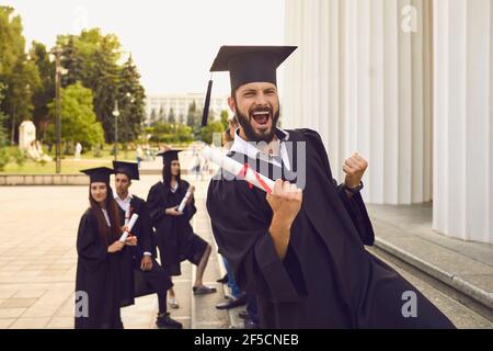 Diplômé émotionnel portant une casquette et une robe de maîtres levant la main avec un diplôme. Banque D'Images