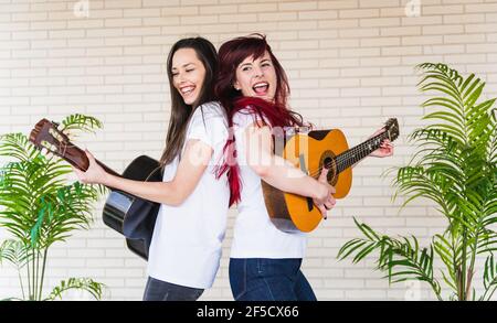 Vue latérale des jeunes femmes souriant et jouant aux guitares espagnoles tout en se tenant dos à dos contre le mur de briques Banque D'Images