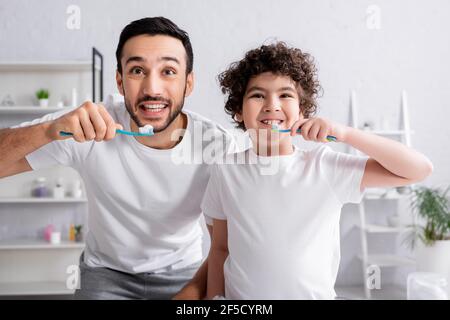 Père et fils musulmans souriants tenant des brosses à dents avec du dentifrice salle de bains Banque D'Images