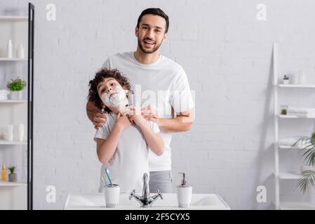 Garçon dans la mousse de rasage tenant le rasoir près du père souriant Banque D'Images