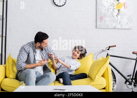 homme arabe pointant avec la main tout en parlant à un fils joyeux sur le canapé à la maison Banque D'Images