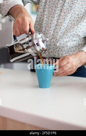 Gros plan sur les mains d'homme senior qui répande une boisson chaude aromatique dans une tasse pendant le petit déjeuner assis dans une cuisine moderne. Grand-père dans la matinée en dégustant un espresso frais brun de la tasse, filtre relax rafraîchissement Banque D'Images