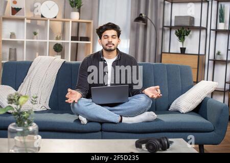 Agréable et sympathique jeune homme indien arabe, assis dans lotus pose avec pc portable sur le canapé dans le salon, et méditant avec les yeux fermés et le sourire, les doigts pliants dans mudra. Banque D'Images