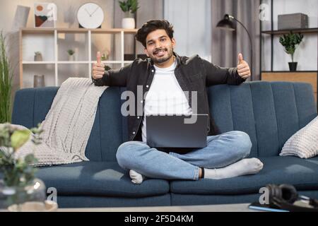 Charmant Indien de 30 ans souriant, assis sur un canapé confortable à la maison, se sentant détendu tout en passant son temps libre avec un ordinateur portable, montrant ses pouces à l'appareil photo. Banque D'Images