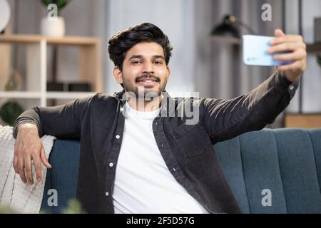 Charmant homme indien souriant, reposant sur un canapé confortable à la maison et utilisant un smartphone pour prendre des photos ou passer un appel vidéo. Loisirs à la maison, concept gadgets Banque D'Images