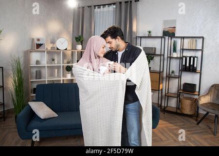 Portrait d'un jeune couple arabe amoureux, ensergeant sous une couverture à la maison et regardant l'un l'autre, touchant les fronts, buvant du café ou du thé. Concept de famille et de relations heureux. Banque D'Images