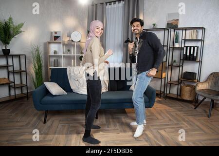 Romantique drôle de conjoints musulmans arabes dansant dans le confortable salon intérieur à la maison, s'amuser et rire, appréciant passer du temps ensemble. Banque D'Images