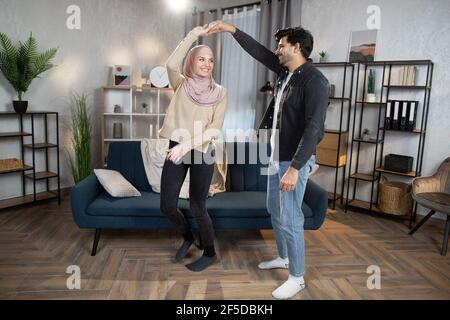 Famille heureuse, jeune couple musulman, appréciant d'être ensemble en dansant à l'intérieur, dans un salon confortable et moderne décoré. Loisirs, détente active. Banque D'Images