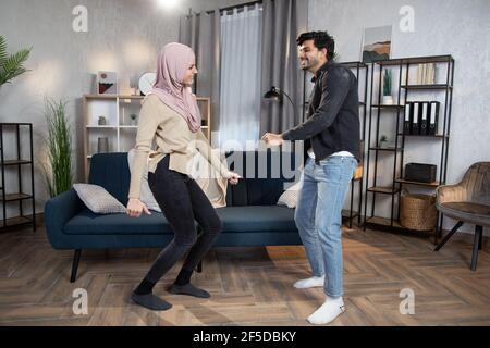 Romantique drôle de conjoints musulmans arabes dansant dans le confortable salon intérieur à la maison, s'amuser et rire, appréciant passer du temps ensemble. Banque D'Images