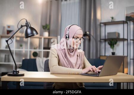 Femme musulmane de 30 ans assez confiante et concentrée dans un hijab avec casque, travaillant sur un ordinateur portable à la maison. Freelance et le concept d'enseignement à distance. Banque D'Images