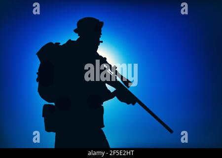 Silhouette du combattant des forces d'élite de l'armée debout avec un fusil de sniper sur fond de ciel bleu avec lune ou soleil. Sniper ou marksman dans un chapeau de boonie, portant un sac à dos en mission, patrouiller la nuit Banque D'Images