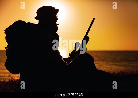 Silhouette de commando combattant, de tireur des forces spéciales de l'armée assis sur la mer ou sur la rive de l'océan au coucher du soleil. Un rifleman de la garde côtière observant la plage, se reposant pendant une patrouille côtière ou une opération amphibie Banque D'Images