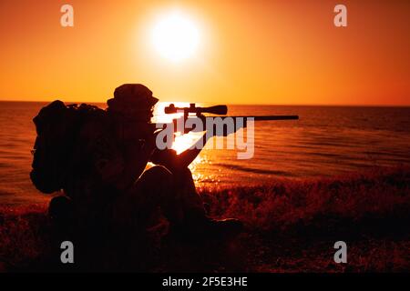 Sniper de l'équipe de commando, tir de tir des forces spéciales de l'armée, tir de fusil de sniper tout en étant assis sur la mer ou sur la côte de l'océan pendant le coucher du soleil. Un soldat de la côte ou du garde-frontière observe la côte avec une vue optique Banque D'Images