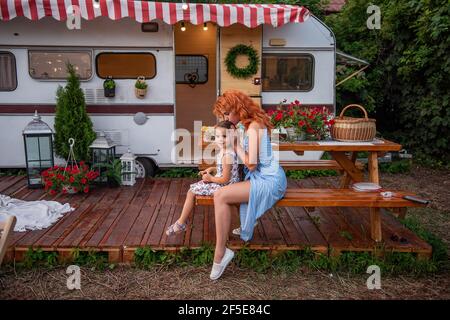 La jeune mère à poil rouge braid les braïdes de sa petite fille sur un banc en bois par camion-remorque. Soins maternels. La jeune fille boit de la limonade. Pique-nique, campin Banque D'Images