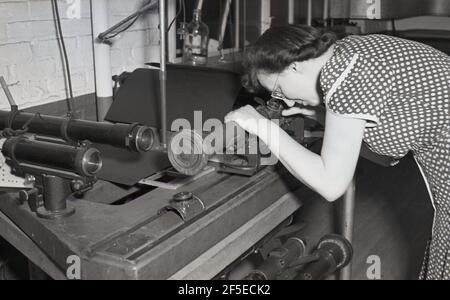 années 1950, historique, une femme porte une robe à pois ou pois dans une usine d'ingénierie légère regardant à travers une petite fenêtre sur une tige métallique en fonte, probablement en vérifiant une lentille de verre. Banque D'Images