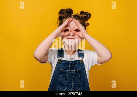 Drôle souriant petite fille d'âge préscolaire faire des doigts comme des lunettes de lunettes forme regardant à travers des jumelles souriantes regardant l'appareil photo Banque D'Images