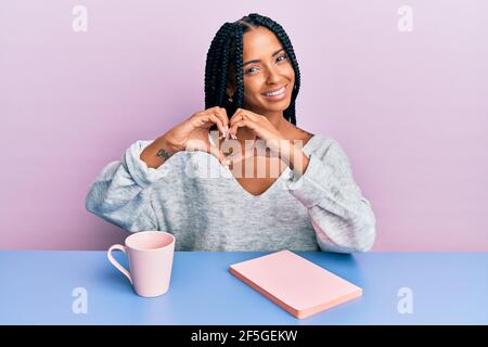 Belle femme hispanique lisant un livre et buvant un café souriant dans l'amour montrant le symbole du coeur et la forme avec les mains. Concept romantique. Banque D'Images