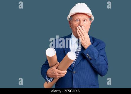 Homme senior beau gris-cheveux portant un casque d'architecte tenant des bleus couvrant la bouche avec la main, choqué et peur d'erreur. Surpris expre Banque D'Images