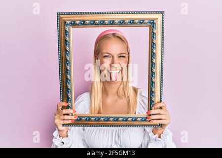 Jeune fille blonde tenant le cadre vide collant la langue dehors heureux avec l'expression drôle. Banque D'Images