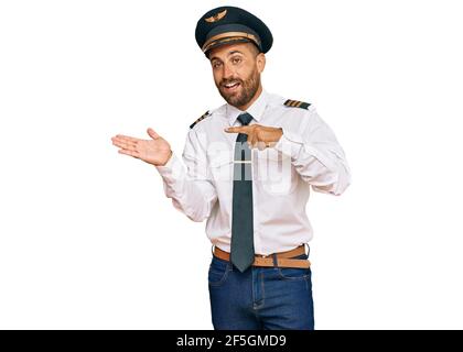 Beau homme avec barbe portant l'uniforme pilote d'avion stupéfié et souriant à l'appareil photo tout en présentant avec la main et en pointant avec le doigt. Banque D'Images