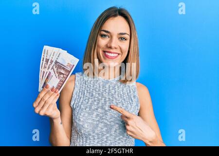 Jeune femme blonde caucasienne tenant des billets en livres égyptiens souriant heureux pointer avec la main et le doigt Banque D'Images