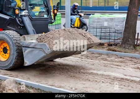 Une petite niveleuse se déplace le long d'un trottoir en construction et porte un seau de sable sur la toile de fond d'une route de la ville. Banque D'Images