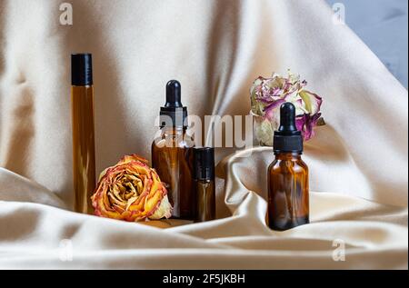 Divers contenants pour le stockage de cosmétiques en verre foncé se trouvent sur les plis de tissu satiné de soie, entouré de fleurs de rose séchées. Parfum, sérum ou Banque D'Images