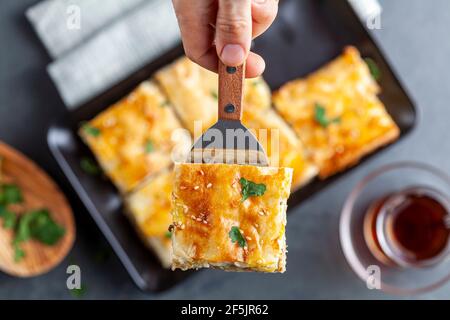 Image de la pose plate montrant une femme caucasienne servant une tranche De kiymali turc borek sur une spatule avec plateau flou De pâtisserie et un verre de thé turc Banque D'Images