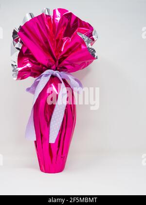 Œuf de Pâques au chocolat géant enveloppé d'une feuille d'argent rose avec un joli noeud à pois violet. Arrière-plan vertical blanc Banque D'Images