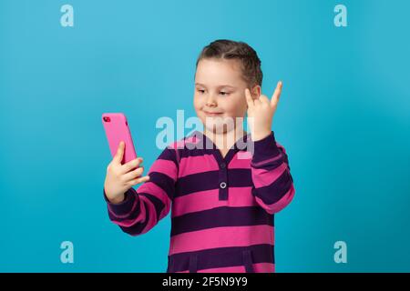 gros plan portrait d'une petite fille souriante avec des queues de porc parlant sur un téléphone de chat vidéo, montrant des signes de cornes avec ses doigts, isolé sur fond bleu Banque D'Images