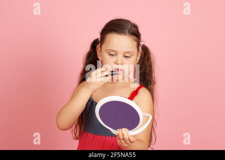 une jeune fille de cinq ans avec des cils peints regarde dans le miroir et peint ses lèvres avec un rouge à lèvres brillant pêche, isolé sur un fond rose Banque D'Images