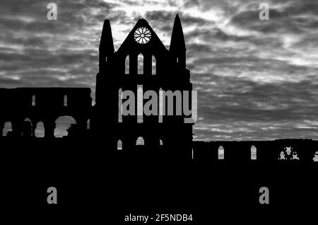 Whitby Abbey, North Yorkshire, Royaume-Uni en silhouette contre un ciel nuageux d'hiver avec le soleil se brisant. B&W. Banque D'Images