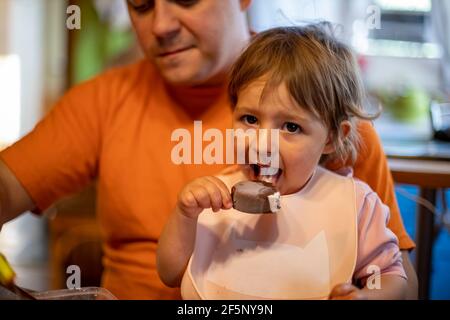 un adorable tout-petit aime manger de la glace à la popsicle sur les genoux de ses parents Banque D'Images