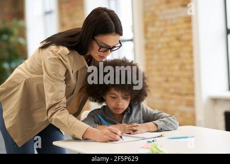 Guidant la jeune femme enseignante dans des lunettes aidant le petit garçon avec la tâche. Enfant étudiant à l'école primaire, assis au bureau Banque D'Images