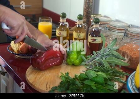 Les mains de l'homme coupant un gros poivron rouge sur une planche de bois avec toutes les herbes autour et les légumes, ingrédients nécessaires pour un repas végétarien Banque D'Images