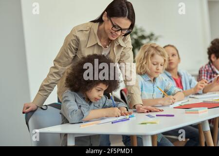 Une jeune femme attentionnée en lunettes aide un petit garçon d'école. Enfants assis à la table, étudiant en classe à l'école primaire Banque D'Images