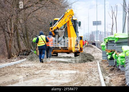 Une pelle hydraulique sur route orange et un groupe de travailleurs préparent une base sablonneuse pour le pavage de dalles sur un sentier de randonnée le long d'une rue de la ville, un après-midi de printemps. Banque D'Images