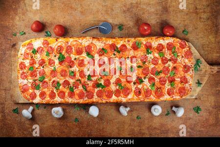 Une longue pizza avec salami et tomates se trouve sur une table en bois. Pizza romaine sur une pelle en bois. Une pizza d'un mètre. Banque D'Images