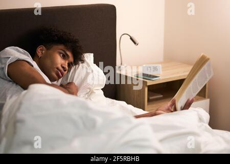 Jeune homme allongé dans le lit à la maison, se reposant et lisant un livre intéressant avant d'aller dormir Banque D'Images