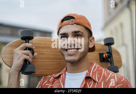 Gros plan portrait d'un jeune patineur tenant une longue planche, marchant dans la rue, souriant. Un Indien heureux portant des vêtements décontractés posant pour les photos Banque D'Images