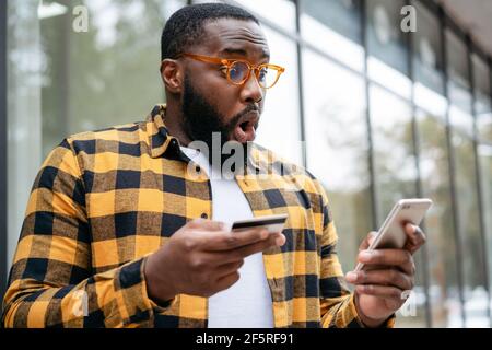 Homme afro-américain enthousiaste tenant une carte de crédit, utilisant l'application mobile pour le shopping en ligne. Un travailleur indépendant émotif reçoit le paiement Banque D'Images