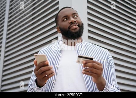 Jeune homme afro-américain élégant tenant une carte de crédit, en utilisant un smartphone pour les achats en ligne. Les jeunes freelance pensive reçoivent le paiement Banque D'Images